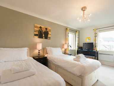 A bedroom with two single beds side by side with white bedding a TV on a black table and two windows at Castle View at the Viking in Seahouses
