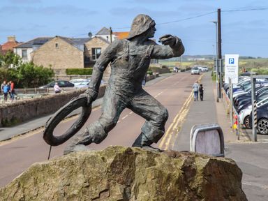 A statue of a man throwing a lifebuoy on a rock by a street with parked cars and buildings in the background at Castle View at the Viking in Seahouses