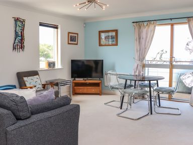 A living room with a gray sofa a glass dining table with four chairs a television on a wooden stand and large windows at Castle View at the Viking in Seahouses