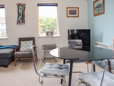 A dining area with a black round table and clear chairs with cushions beside a sitting area with a gray armchair and a radio on a window sill at Castle View at the Viking in Seahouses
