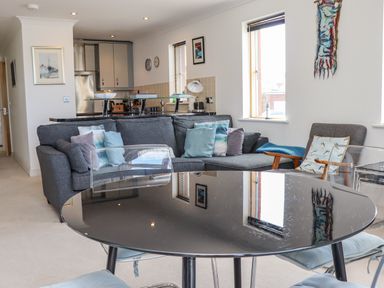 A living room with a gray sofa with pillows a black round table with transparent chairs and a kitchen in the background at Castle View at the Viking in Seahouses