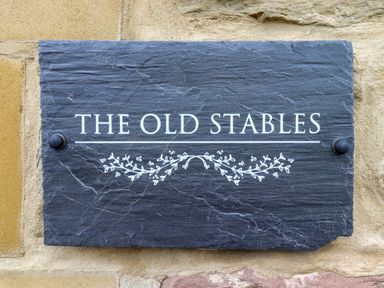 A black stone plaque with the text the old stables and a decorative floral design on a stone wall at The Old Stables in Bamburgh