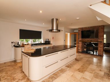 A kitchen with a large island, stovetop, and a chimney hood with a brick fireplace and mounted TV in the adjoining living area at The Old Stables in Bamburgh