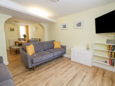 A living room with a gray sofa with yellow pillows a wall-mounted TV and a bookshelf with books and a dining area in the background at Cove Street Cottage Weymouth