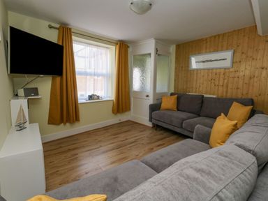A living room with two gray sofas with yellow pillows a wall-mounted TV and a window with yellow curtains at Cove Street Cottage Weymouth