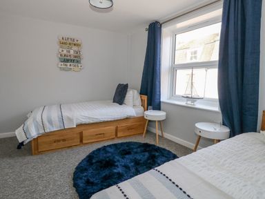 A bedroom with two single beds two white round bedside tables a window with blue curtains and a decorative sailboat on the windowsill at Cove Street Cottage in Weymouth