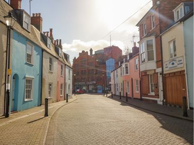 A street with colorful terraced houses and a large brick building in the background at Cove Street Cottage in Weymouth