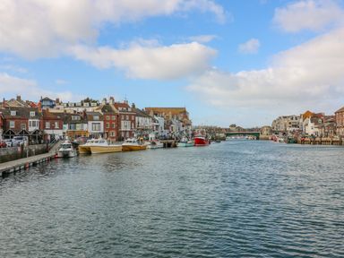 A harbor with boats docked along both sides and buildings lining the waterfront at Cove Street Cottage Weymouth