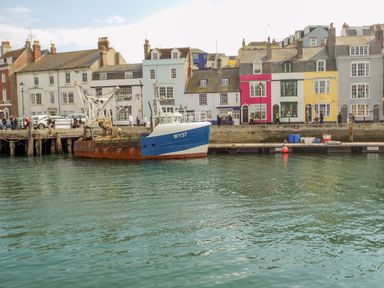 A fishing boat moored at a harbor with colorful buildings and people walking along the waterfront at Cove Street Cottage in Weymouth