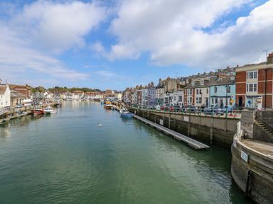A river with boats docked alongside buildings and a street with cars at Cove Street Cottage in Weymouth