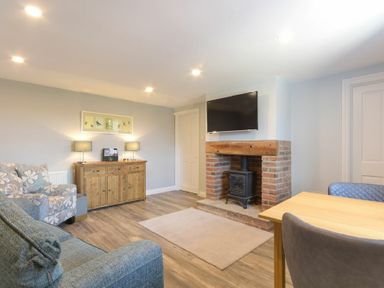 A living room with a brick fireplace and TV above it a wooden sideboard with lamps a floral armchair and a wooden dining table with chairs at Dairymaid's Cottage in Amble