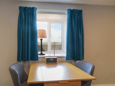 A small wooden table between two gray chairs in front of a window with blue curtains and a lamp and radio on the windowsill at Dairymaid's Cottage in Amble