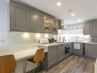 A kitchen with gray cabinets wooden chairs and a window at Dairymaid's Cottage in Amble