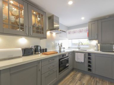 A kitchen with grey cabinets a built in oven countertop appliances and a window at Dairymaid's Cottage in Amble