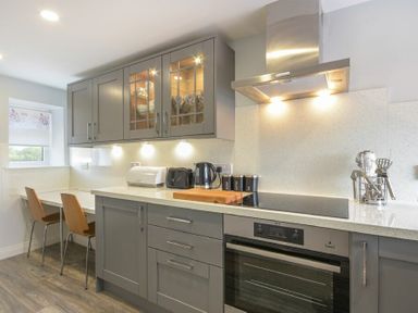 A kitchen with grey cabinets a built in oven a stovetop a range hood a wooden cutting board a kettle toaster bread bin and two chairs at Dairymaids Cottage in Amble