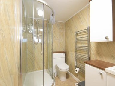 A bathroom with a curved glass shower enclosure a white toilet chrome towel rack and white cabinets at Dairymaids Cottage in Amble