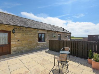 An outdoor patio with two chairs and a small round table next to a stone building with two windows and a wooden door at Dairymaid's Cottage in Amble