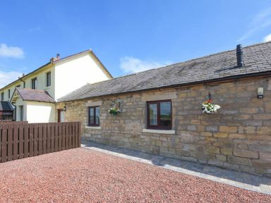 A stone exterior wall with two windows and hanging flower baskets outside a cottage with a gravel front yard at Dairymaid's Cottage in Amble