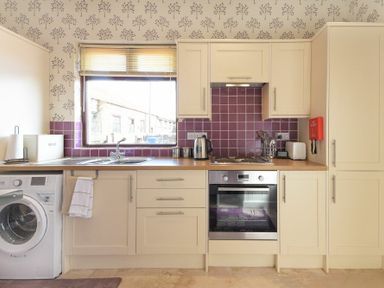 A kitchen with cream cabinets purple tile backsplash and a washing machine under the counter at Dairyman's Cottage in Amble