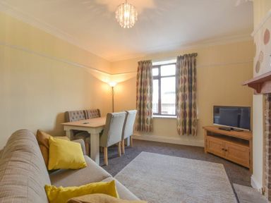A living room with a sofa yellow cushions dining table chairs window curtains television on wooden stand at Dairyman's Cottage in Amble