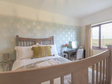 A bedroom with a wooden bed frame, bedside tables with lamps, and a desk with a chair and flowers at Dairyman's Cottage in Amble