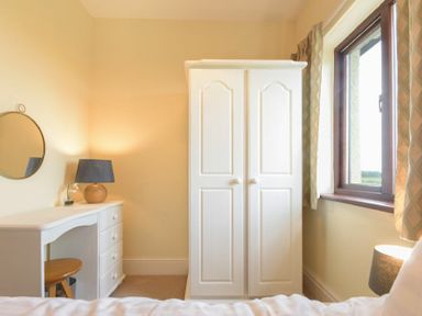 A bedroom corner with a white wardrobe a desk with a lamp and stool and a window with curtains at Dairyman's Cottage in Amble