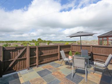 An outdoor patio with stone tiles a table with an umbrella and four chairs surrounded by a wooden fence at Dairyman's Cottage in Amble