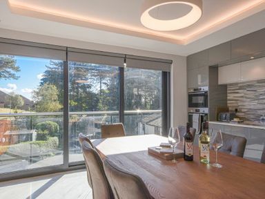 A dining area with a wooden table, six chairs, wine bottles, wine glasses, and large windows overlooking trees at Riverview (Alnmouth) in Alnmouth