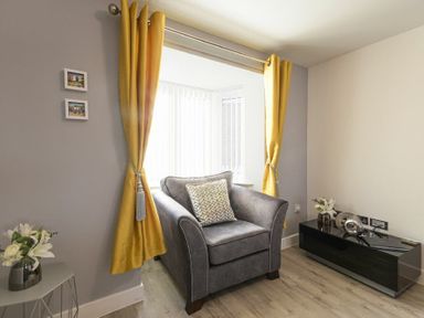 A sitting area with a gray armchair yellow curtains side table and black cabinet with decorations