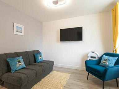 A living room with a dark sofa with blue bee pillows a blue armchair with a matching pillow a wall-mounted TV and yellow curtains at Coastal View in Seahouses
