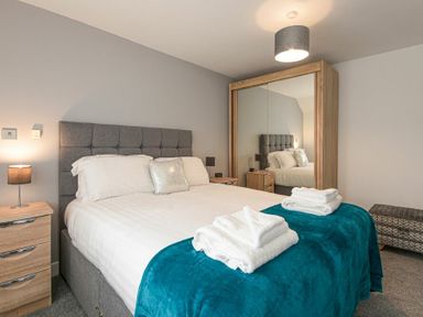 A bedroom with a double bed with white linens and teal blanket bedside tables with lamps and a mirrored wardrobe at Coastal View in Seahouses