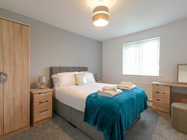 A bedroom with a double bed with white bedding and a teal throw a wardrobe bedside tables with lamps and a dressing table at Coastal View in Seahouses