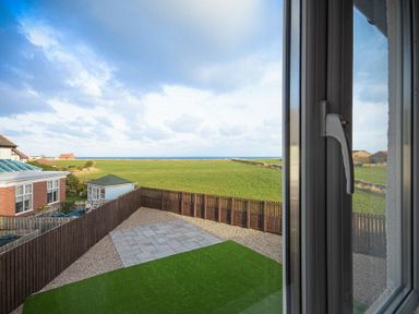 A fenced backyard with a patio and grass area overlooking green fields and the sea at Coastal View in Seahouses