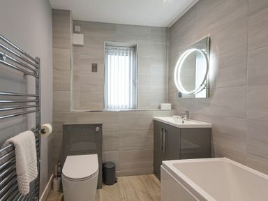A bathroom with a toilet sink under a lit circular mirror and a bathtub at Coastal View in Seahouses