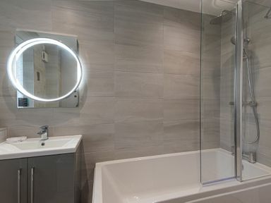 A bathroom with a sink and cabinet a round illuminated mirror a bathtub with a glass shower screen and a shower at Coastal View in Seahouses