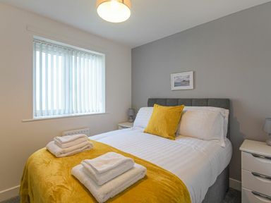 A bedroom with a bed with white and yellow bedding two bedside tables and a window with vertical blinds at Coastal View in Seahouses
