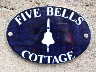 An oval dark blue sign with white text reading Five Bells Cottage mounted on a stone wall