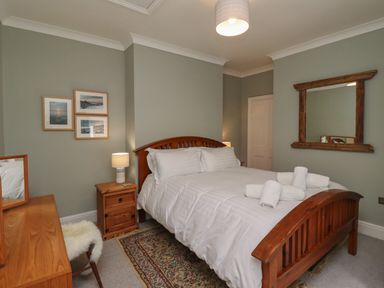 A bedroom with a wooden bed with white linens and rolled towels a wooden nightstand with a lamp framed pictures on the wall a wooden dresser with a mirror and a chair with a white fur cover a patterned rug on the floor at Five Bells Cottage in Alnmouth