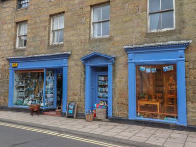 A street view of a stone building with two shop windows and a door painted blue displaying various items at Five Bells Cottage in Alnmouth