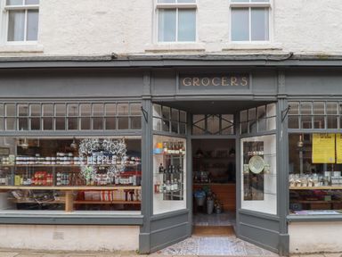 The exterior of a grocers shop with large display windows showing shelves stocked with jars and bottles at Five Bells Cottage in Alnmouth