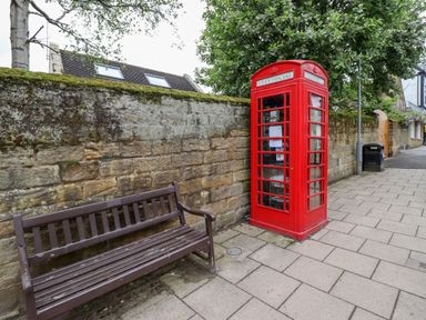 A red telephone booth next to a brown bench by a stone wall on a sidewalk at Five Bells Cottage in Alnmouth