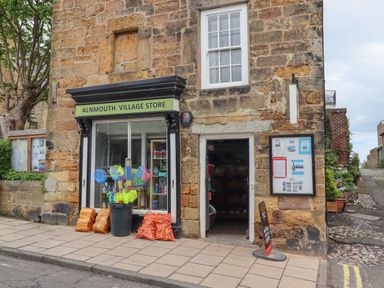 The exterior of Alnmouth Village Store showing a stone building with a display window, entrance door, and items like nets and firewood outside on the sidewalk at Five Bells Cottage in Alnmouth