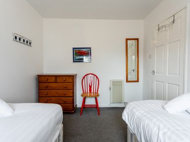 A bedroom with two beds a wooden chest of drawers a red chair a wall mirror and a white door at Palmers Rest in Beadnell
