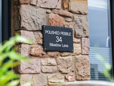 A stone wall with a black sign reading polished pebble 34 meadow lane with a window nearby at Polished Pebble in Beadnell