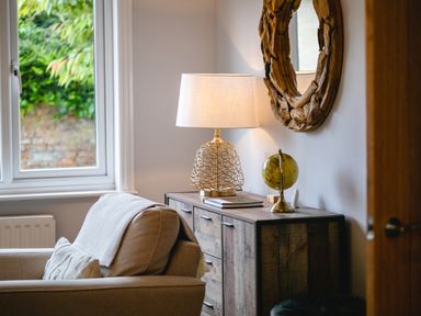 A wooden cabinet with a table lamp and globe beside a beige armchair in a room with a window and a round wooden mirror on the wall at Polished Pebble in Beadnell