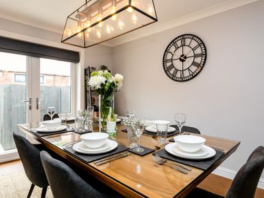 A dining room with a wooden table set for six with white dishes and glasses a large clock on the wall and glass doors at Polished Pebble in Beadnell