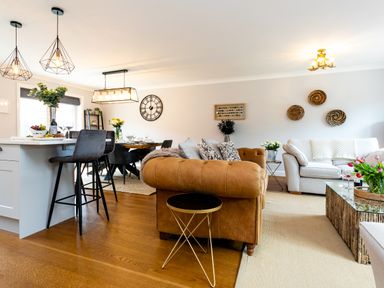 An open plan living and dining area with a brown sofa white chairs a dining table and bar stools at Polished Pebble in Beadnell