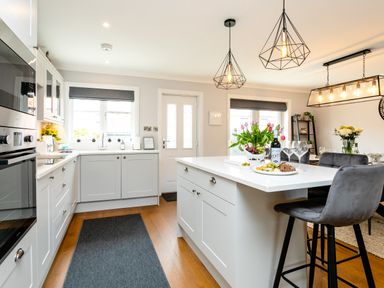 A kitchen with white cabinets island with flowers wine and snacks with dining table and chairs at Polished Pebble in Beadnell