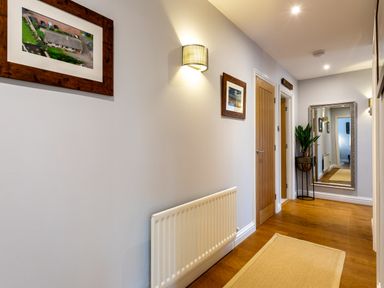 A hallway with wood floor multiple doors framed pictures a wall light a plant in a pot and a large mirror at Polished Pebble in Beadnell
