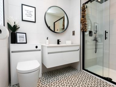 A bathroom with a toilet, a white floating vanity with black faucet and round mirror, a framed picture, and a glass shower at Polished Pebble in Beadnell